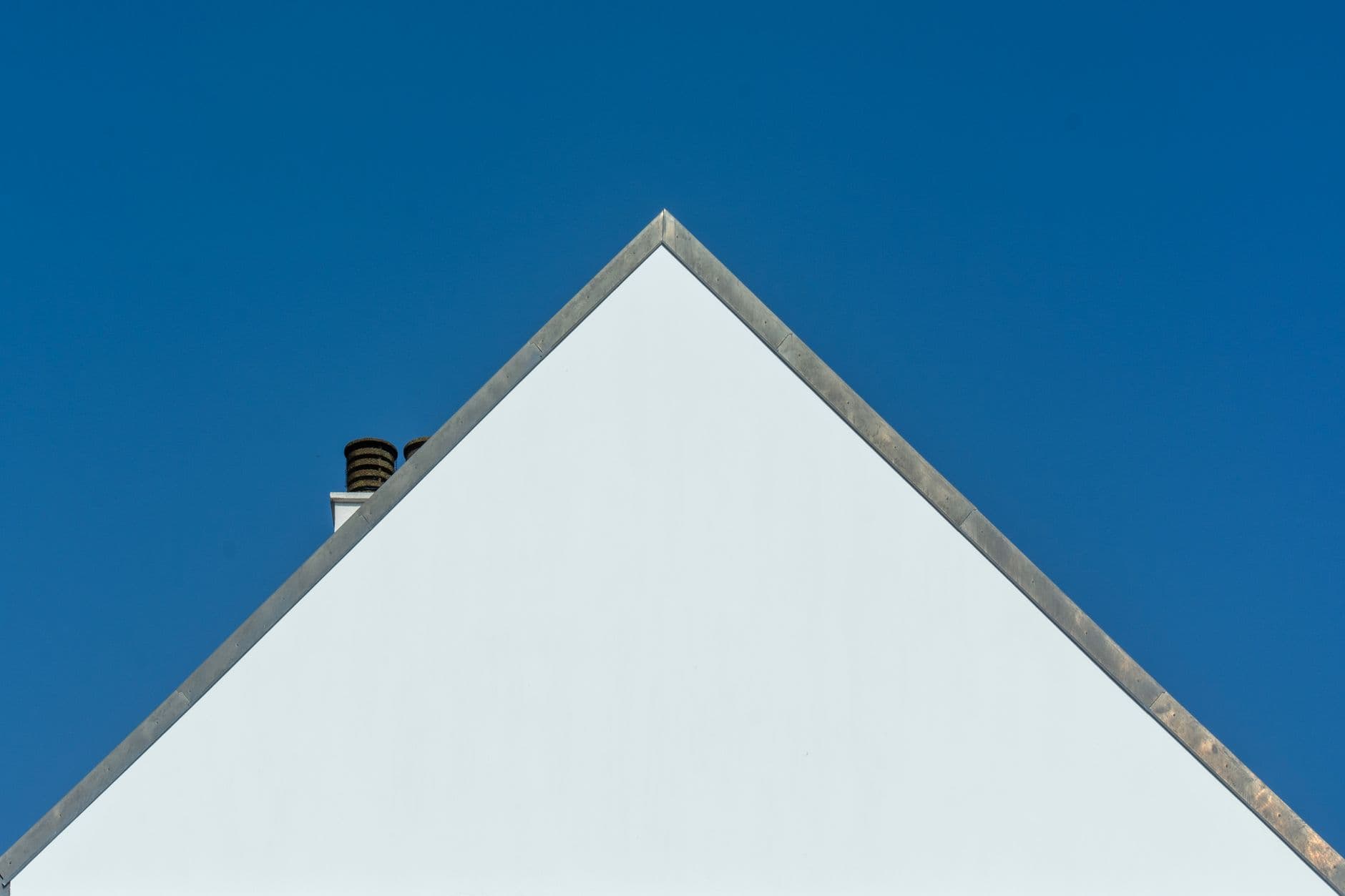 Triangle gable of a white house with chimney against a clear blue sky, highlighting minimalist architecture.